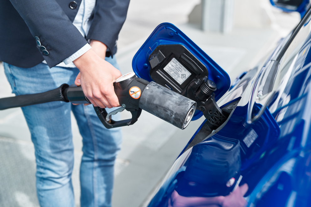 Blaues Auto mit offenem Tankdeckel wird durch Mann mit Wasserstoff betankt.