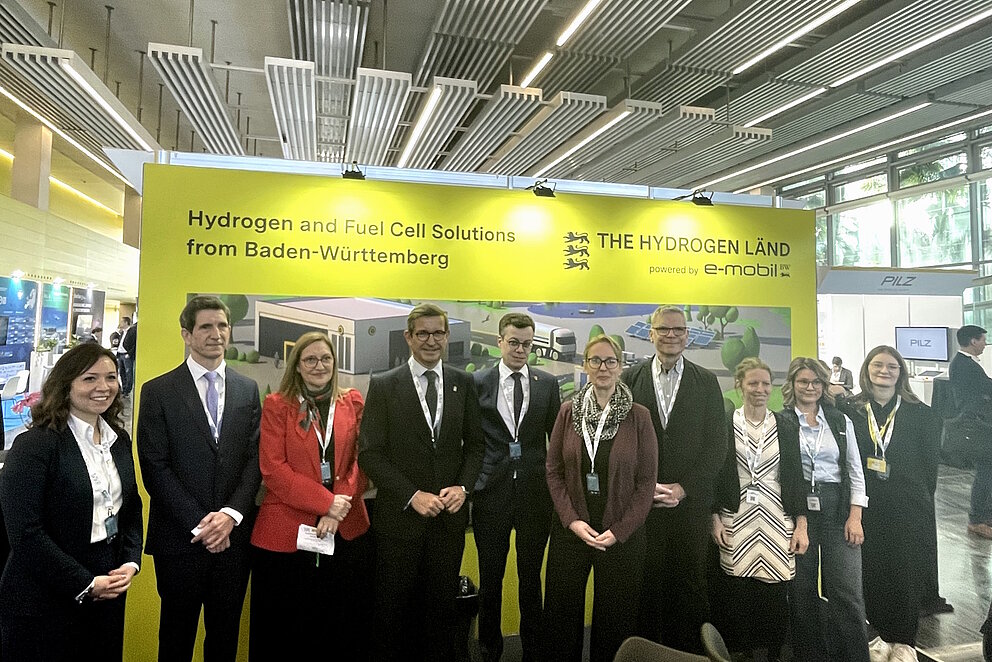 Vor einer gelben Wand mit dem Logo von e-mobil BW posiert eine Gruppe von schick angezogenen Menschen in der Messehalle.