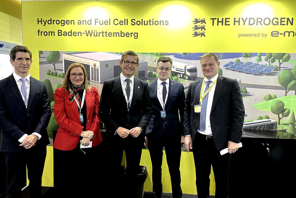 Vor gelber Messewand der e-mobil BW lächeln Regoerungsvertreter Andalusiens mit baden-württembergischen Vertretern der Landespolitik in die Kamera.