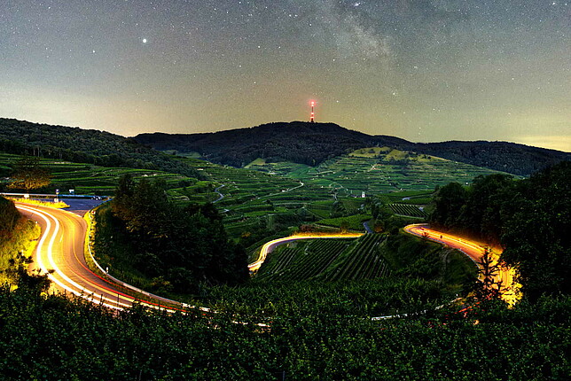 Eine kurvieg Straße windet sich durch bewaldetes Gebiet bei Nacht und es sind Scheinwerfer fahrende Autos erkennbar.