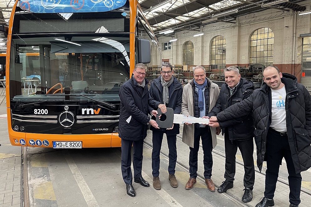 Eine gruppe Männder steht vor Wasserstoffbus in einer Halle und halten einen überdimensionierten Schlüssel in den Händen.