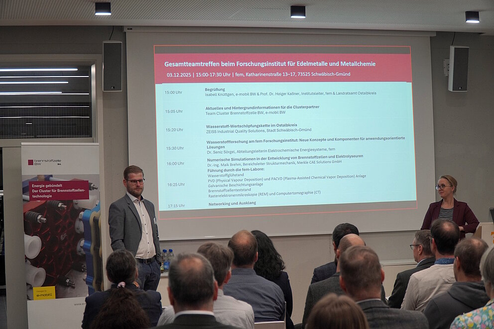 EIne Gruppe Menschen ist von Hinten zu sehen, wie sie sitzend nach Vorne auf eine Präsentation von zwei Referenten blicken.