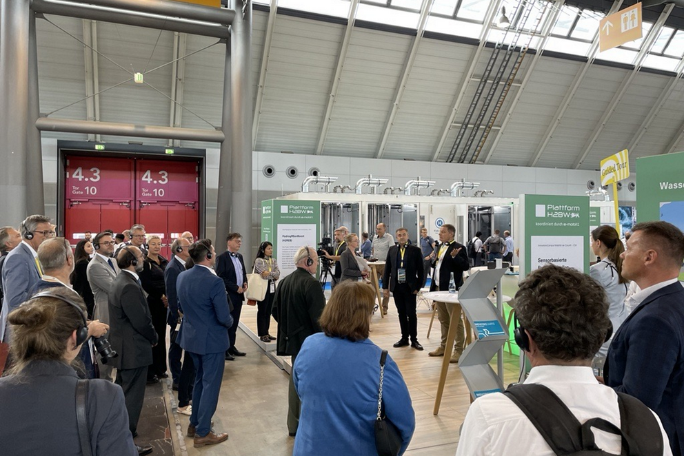 In einer Messehalle steht eine große Gruppe Menschen um den Stand der Plattform H2BW und blickt auf den Stand.