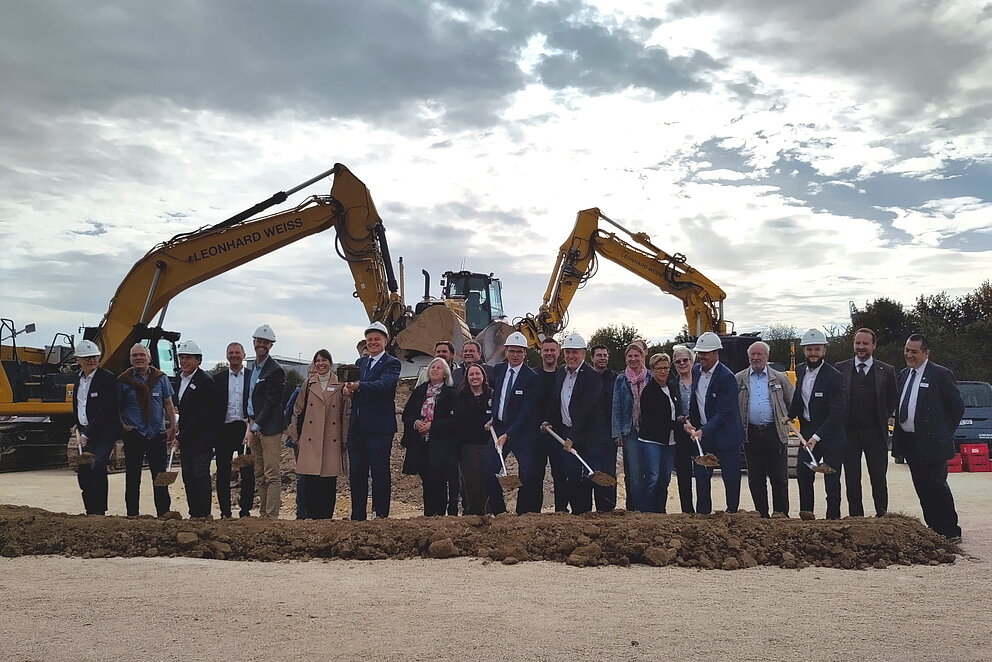 Menschgruppe steht vor zwei Baggern auf der Baustelle der Elektrolyseurs in der Erde.