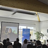 Eine Person spricht per Videokonferenz zugeschalten zum Publikum.