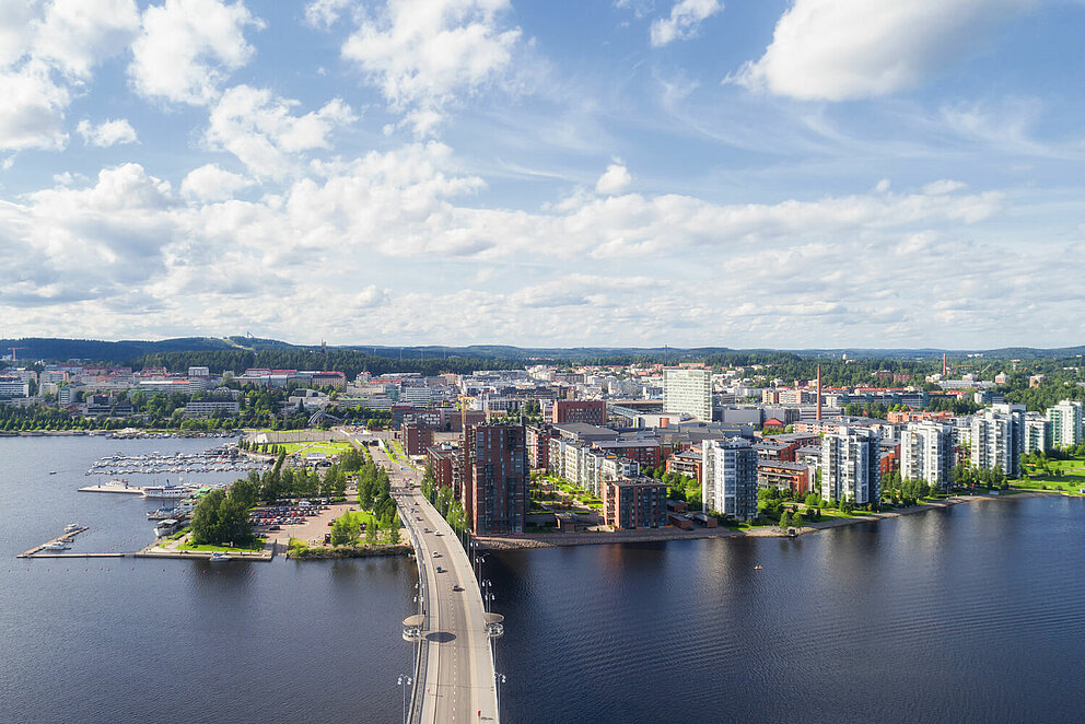 Luftaufnahme einer Stadt am Ufer eines Gewässer, zu dem eine moderne Brücke führt.