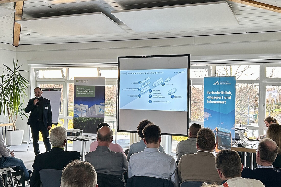 Vor einer Präsentationswand steht ein Referent und spricht vor Zuschauern in ein Mikrophon.