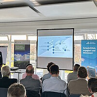 Vor einer Präsentationswand steht ein Referent und spricht vor Zuschauern in ein Mikrophon.