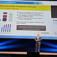 Referent steht auf einer großen Bühne vor einer Leinwand und refereriert.