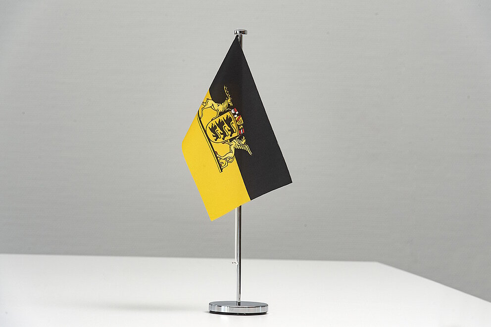 Kleine Flagge des Landes Baden-Württemberg ist an Ständer befestigt und steht vor weißem Hintergrund.
