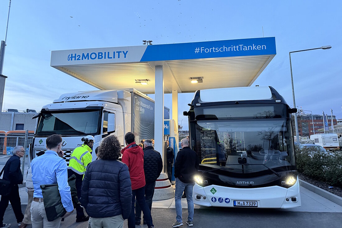 An einer Wasserstoff-Tankstelle stehen nebeneinander ein Lkw und ein Bus und werden vor Zuschauern betankt.