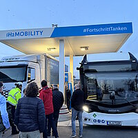 An einer Wasserstoff-Tankstelle stehen nebeneinander ein Lkw und ein Bus und werden vor Zuschauern betankt.