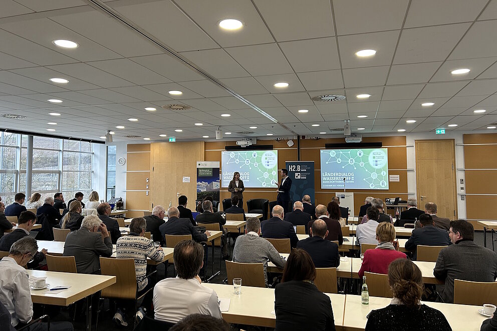 Rund 100 Personen sitzen in einem Raum an Tischen und hören einem Vortrag zu.