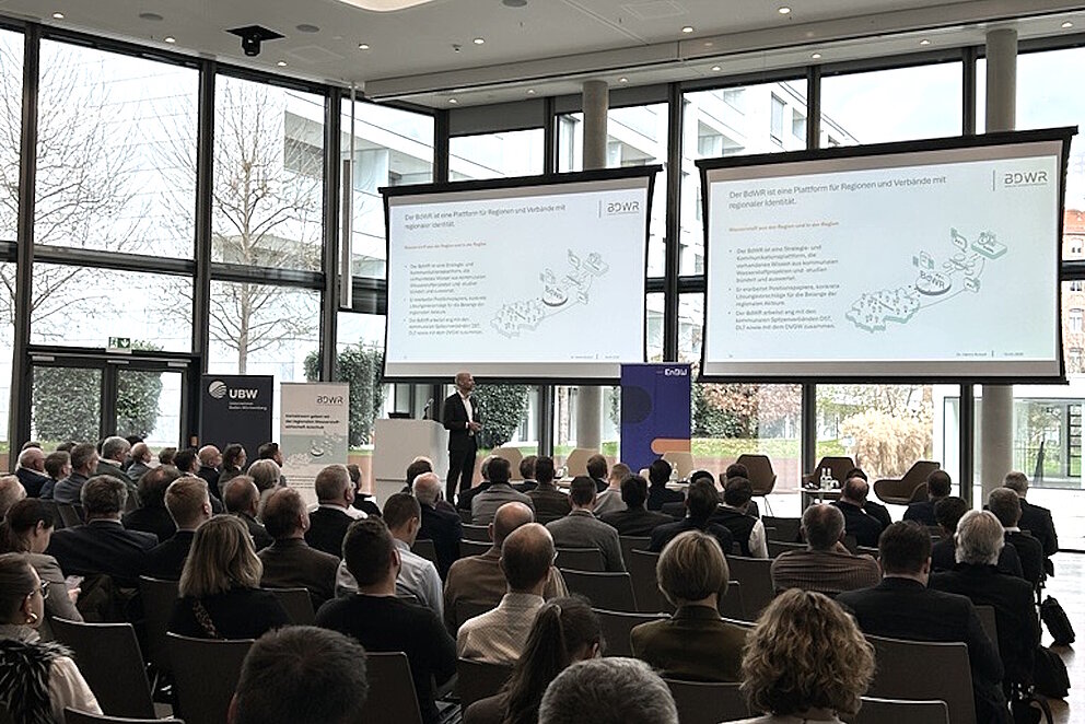 In einem großen Saal sitzen rund 150 Personen und schauen auf zwei Leinwände.