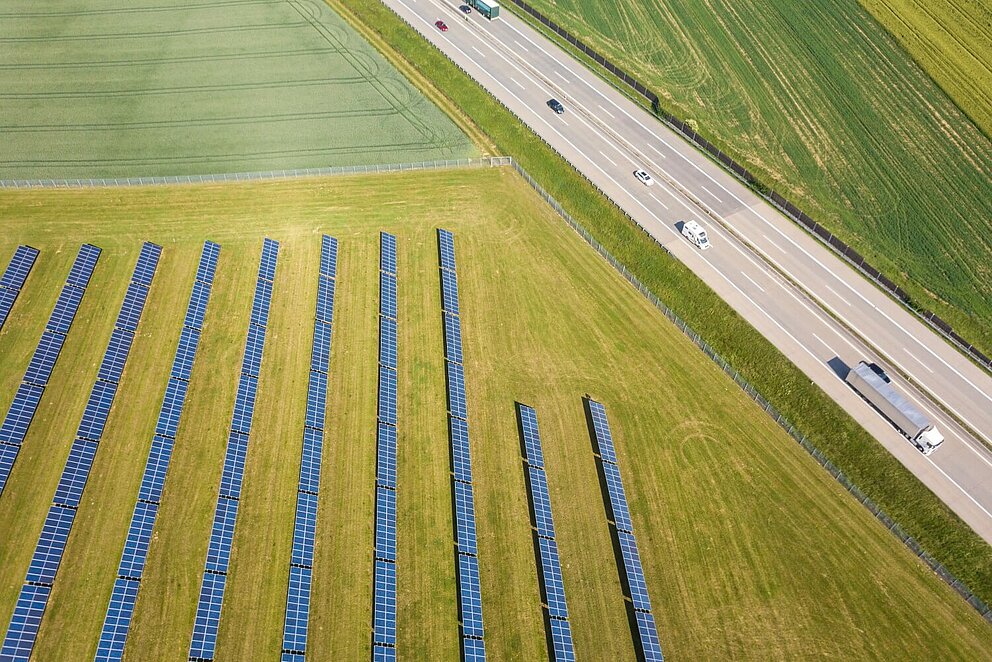 Luftaufnahme einer Photovoltaikanlage auf einer Wiesenfläche neben einer Straße.