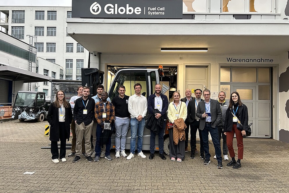 Eine Gruppe Menschen posiert vor dem Eingang einer Lagerhalle mit Firmenlogo.