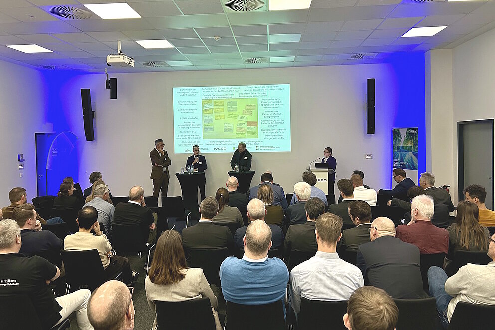 Vier Personen sprechen als Panel vor einer Gruppe von Rund 80 sitzenden Personen.