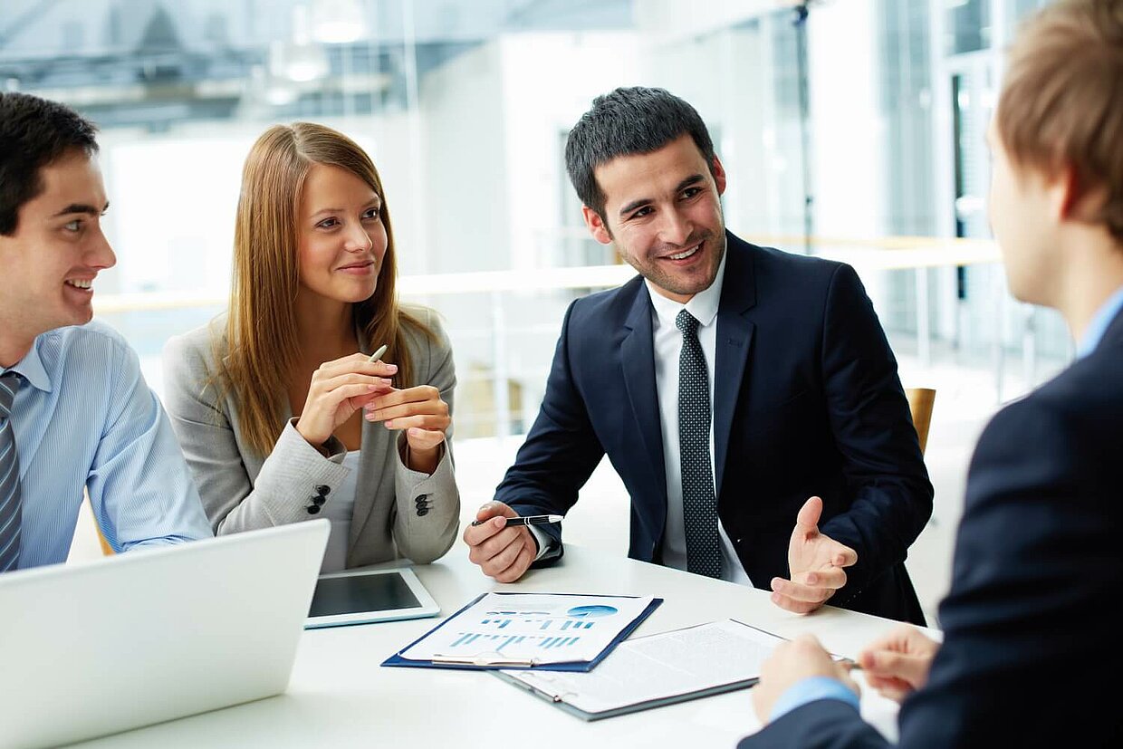 Vier Personen in Business-Kleidung sitzen eng an einem Tisch und unterhalten sich über Unterlagen, die neben einem Laptop auf dem Tisch liegen.