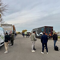 Zwischen aufgereihten Lkw mit H2-Antrieb stehen Gruppen vorn Personen und unterhalten sich auf freier Fläche.