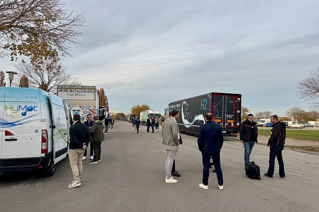 Zwischen aufgereihten Lkw mit H2-Antrieb stehen Gruppen vorn Personen und unterhalten sich auf freier Fläche.