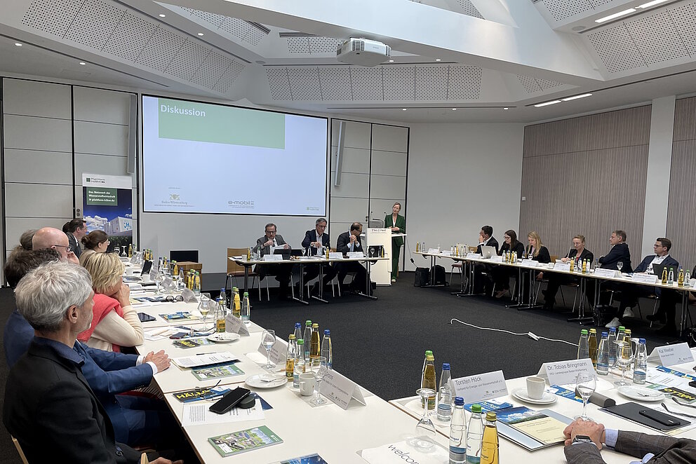 Etwa 30 Personen sitzen an weißen Tischen, die in einem Quadrat angeordnet sind und Blicken zur Stirnseite, wo die Umweltministerin Thekla Walker am Rednerpult spricht.