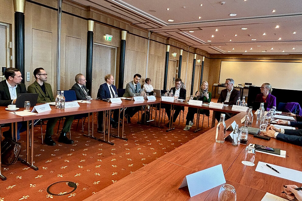 Rund 25 Personen sitzen Tischen, die zu einem U gestellt sind und diskutieren.