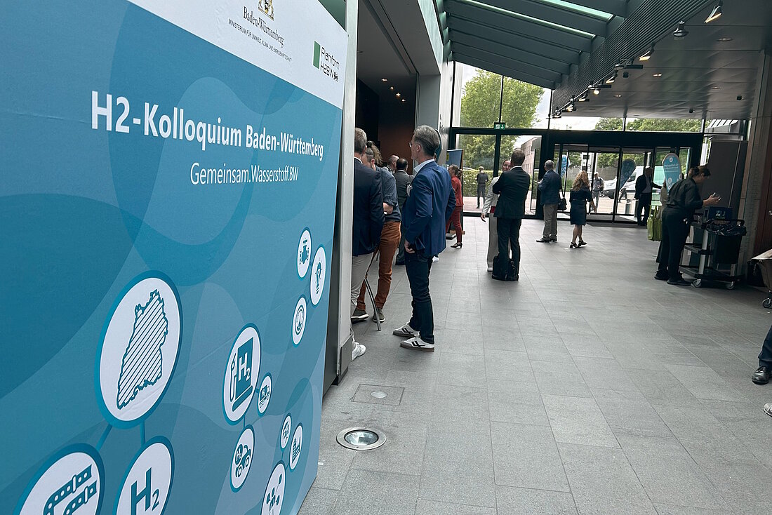 Druck mit großer Aufschrift "H2-Kolloquium" im Vordergrund, im Hintergrund stehen Personen im EIngangsbereich.