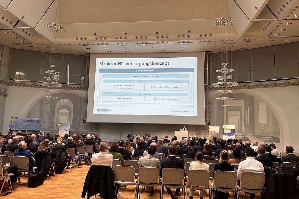 Rund 200 Personen sitzen in Stuhlreihen in einem großen Saal und blicken nach Vorne, wo großflächig eine Präsentation gezeigt wird.