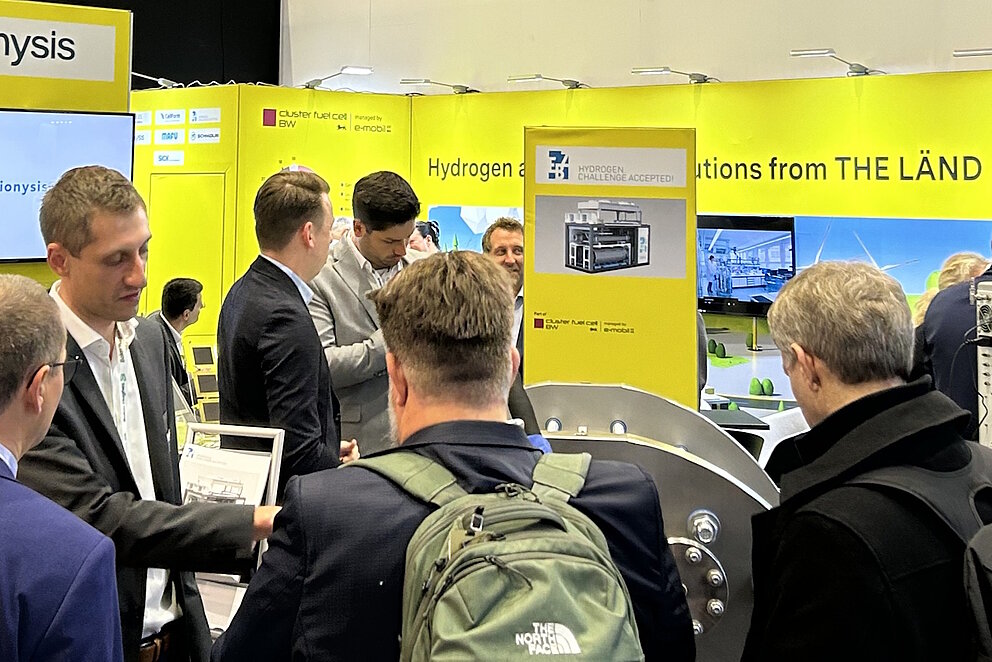 Eine Gruppe Menschen steht auf einem gelben Messestand und lässt sich etwas erklären.