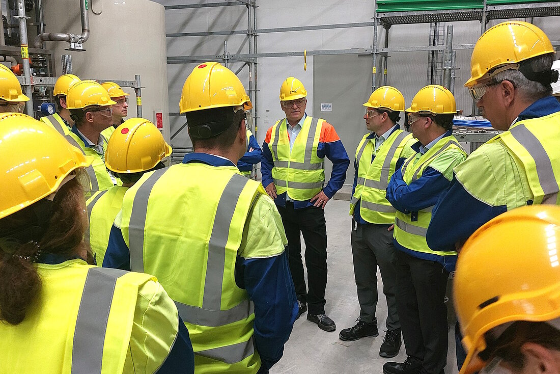 Eine Gruppe steht im Halbkreis in einer Werkshalle und hört dem Leiter zu, der einen Vortrag hält. Alle Tragen gelbe Helme und Warnwesten 