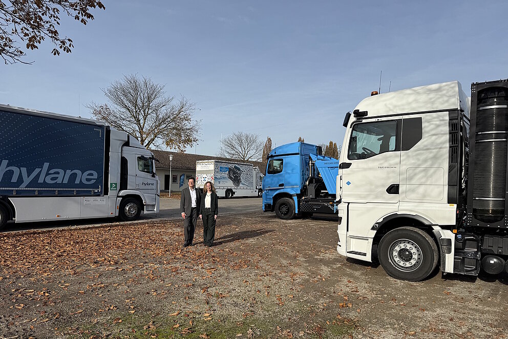 Zwischen drei großen Lkw stehen zwei Personen auf freier Fläche.