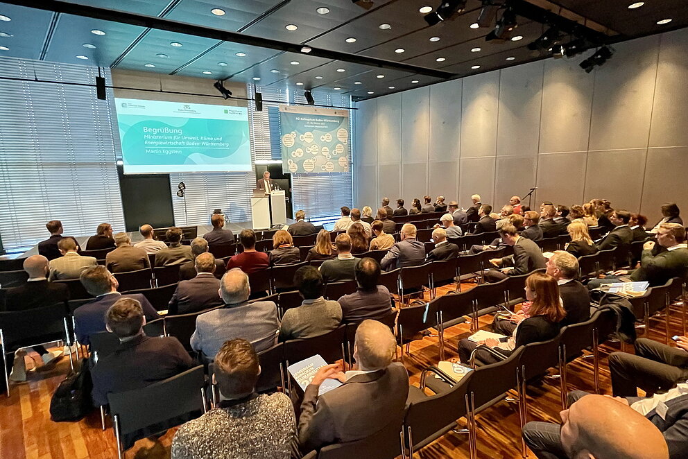 Konferenzraum mit Publikum in Sitzreihen, alle Blicken zur Bühne wo eine Präsentation gezeigt wird.