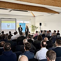 Zwei Referenten stehen vor einer Gruppe von rund 100 Personen und sprechen in ein Mikrophon.