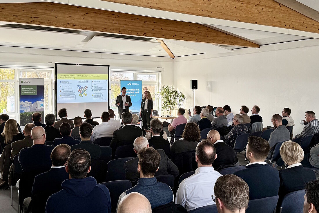 Zwei Referenten stehen vor einer Gruppe von rund 100 Personen und sprechen in ein Mikrophon.