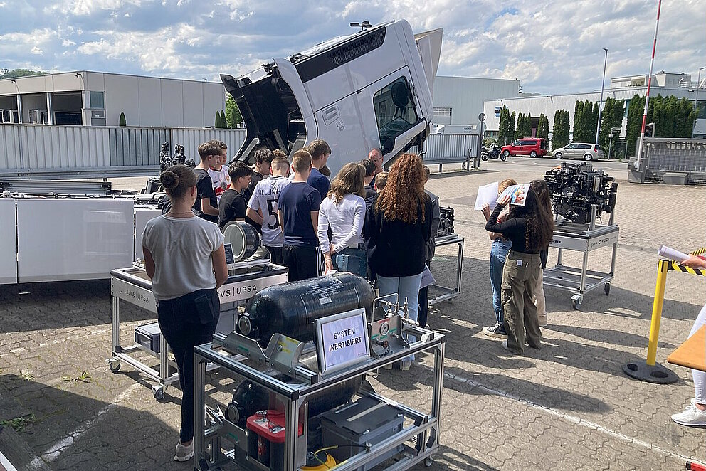 Rund 10 Schüler:innen betrachten ein Exponat einer Brennstoffzelle vor einem mit Wasserstoff betriebenen Lkw. Dieser ist aufgeklappt, d.h. man kann die verbauten Antriebskomponenten sehen.