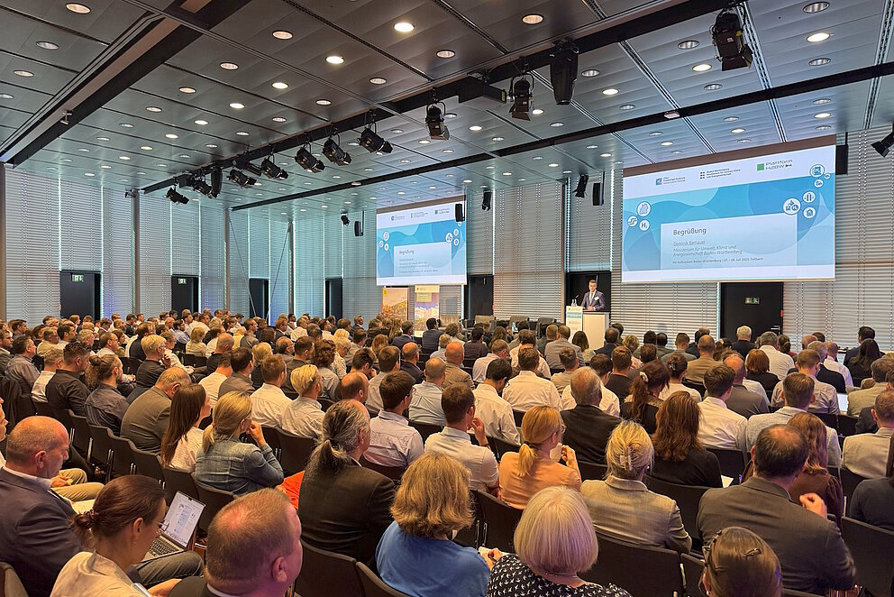 Rund 300 Personen sitzen in Reihen in einem großen Saal und blicken nach Vorn, wo ein Redner steht.