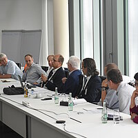 Rund 12 Personen sitzen in einem Bürogebäude an einer langen Tischreihe und disktueiren.