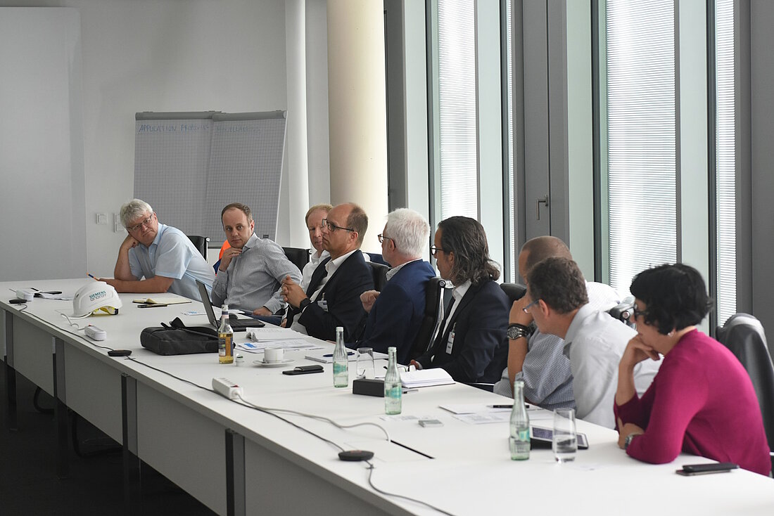 Rund 12 Personen sitzen in einem Bürogebäude an einer langen Tischreihe und disktueiren.