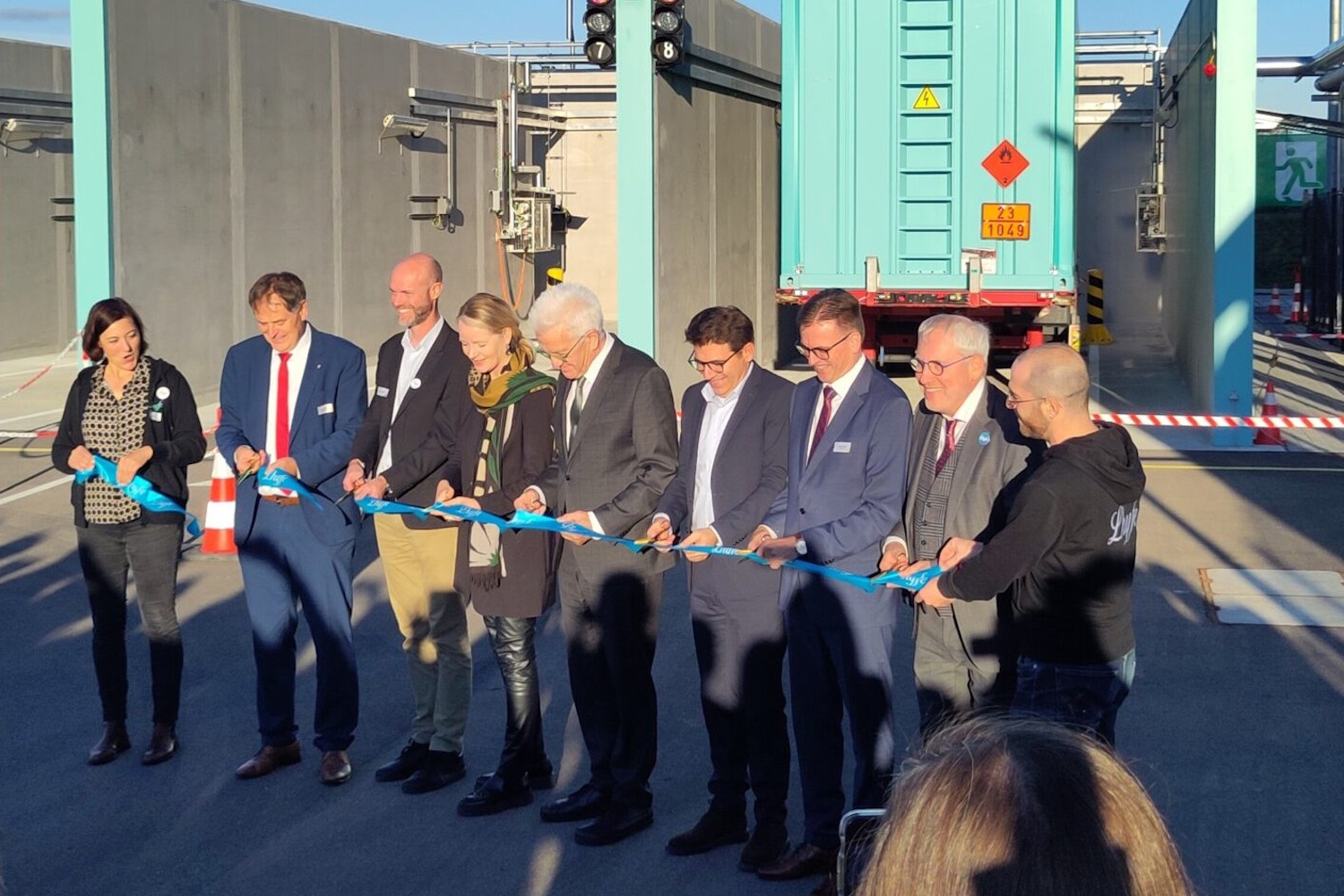 Eine Gruppe Personen steht auf einem Industriegelände und schneidet gemeinsam ein blaues Band durch.