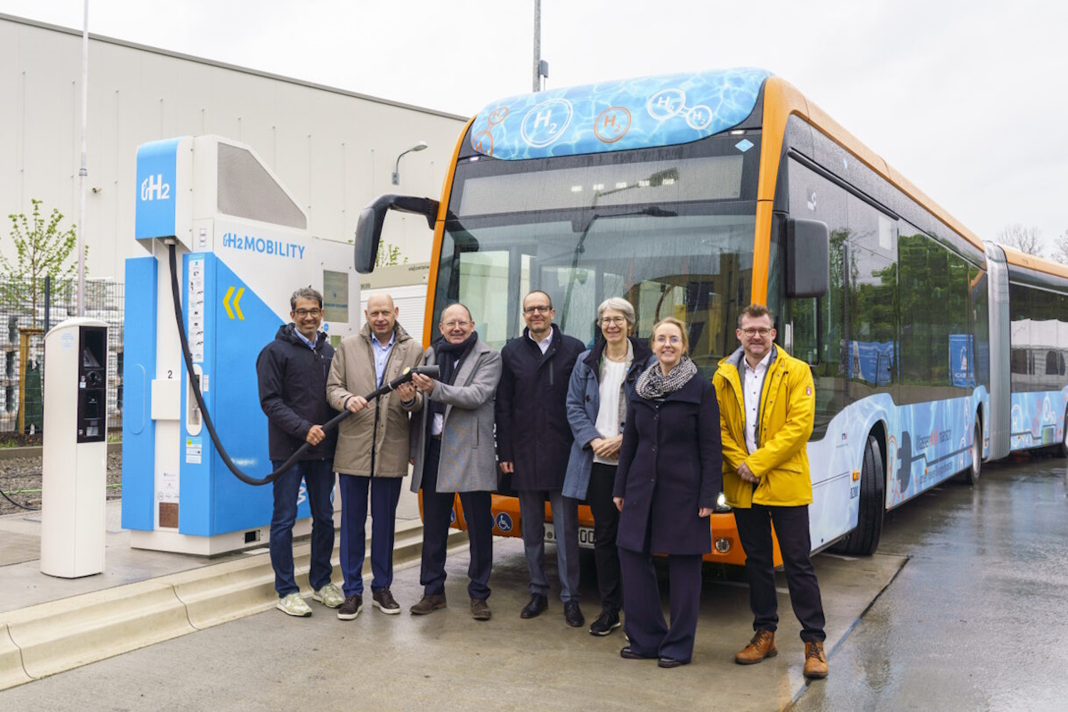 Ein Wasserstoffbus steht an einer blau-weißen Wasserstoff-Tankstelle. Davor haben sich sieben Personen aufgereiht und schauen in die Kamera, darunter der Staatssekretär des Umweltministeriums Dr. Andre Baumann.