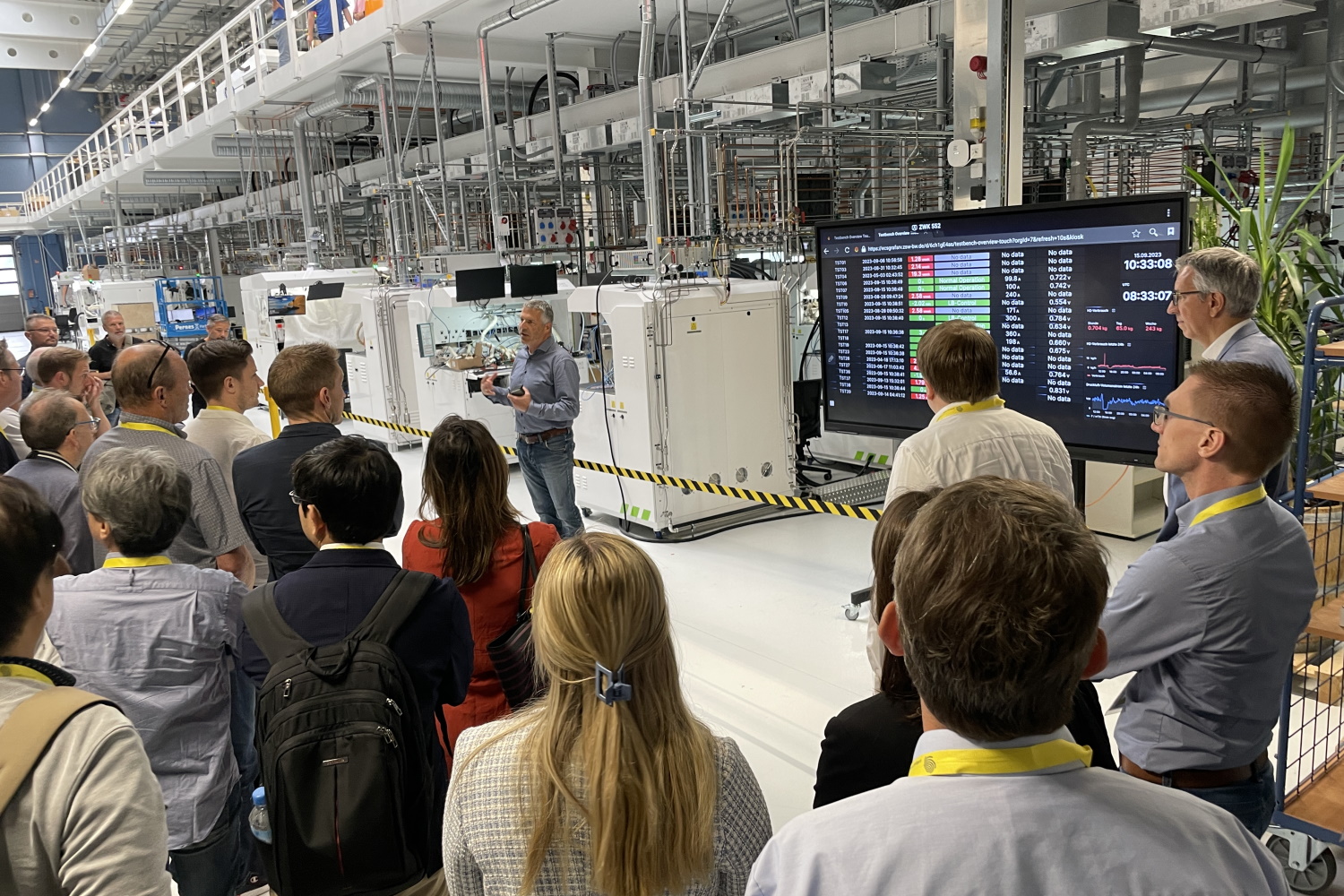 Eine Gruppe Besucher:innen steht im Halbkreis in der Fabrik voller Leitungen und Rohre und blickt auf einen Bildschirm mit technischen Rechnungen.