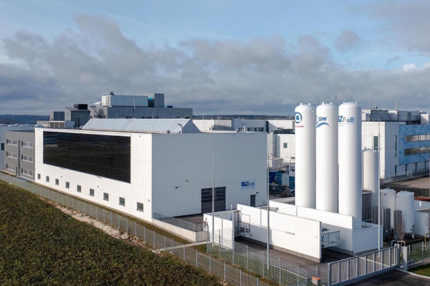Luftaufnahme einer quadratischen, weißen Fabrik mit großem Photovoltaik-Modul, das die ganze Außenwand füllt.