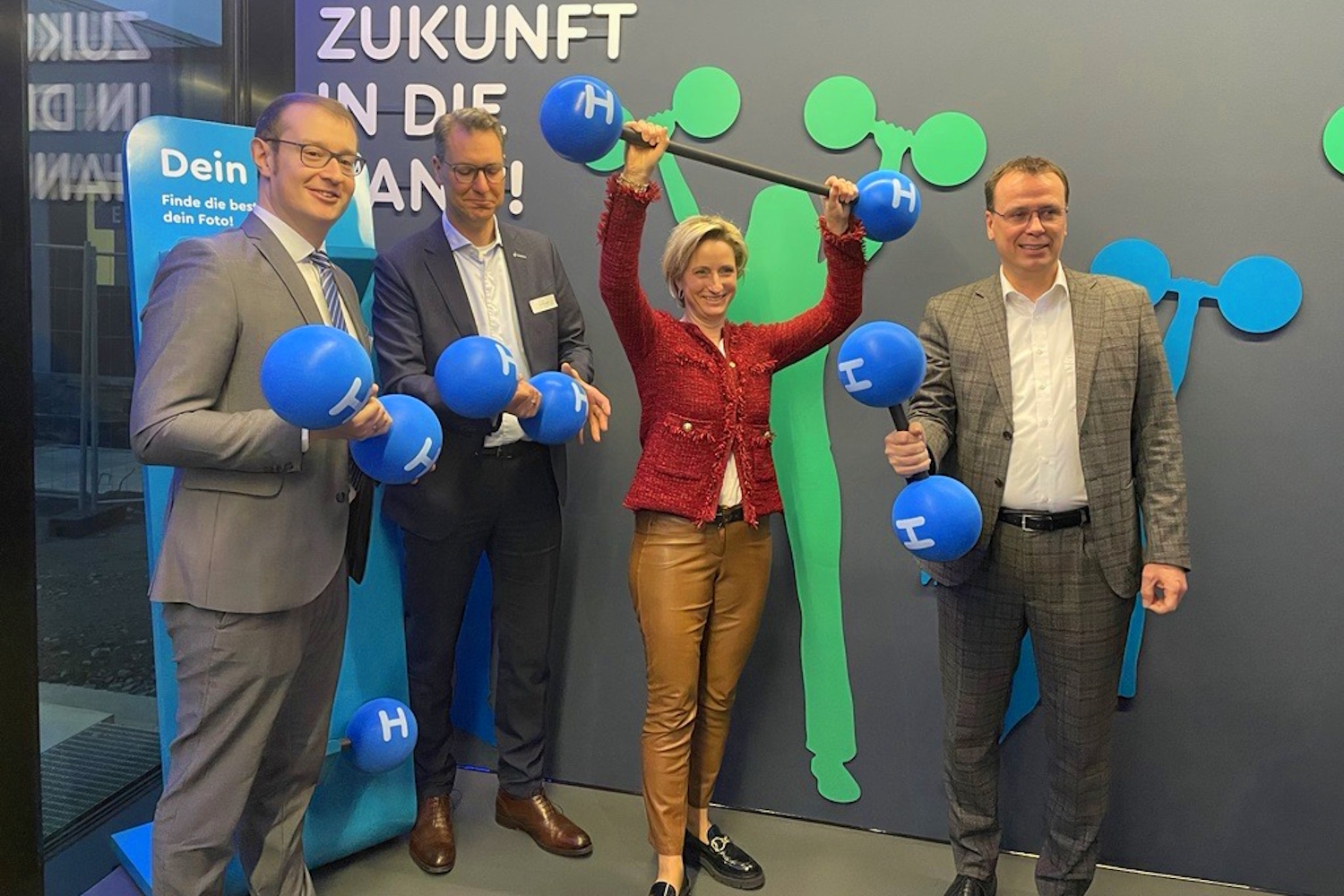 Die Wirtschaftminsiterin und weitere Abgeordnete posieren vor einem Schriftzug das Wort Zukunft enthält und halten Modelle von Wasserstoffmolekülen hoch.