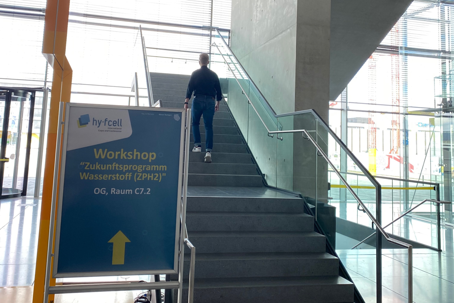 Ein Schild weist den Weg zum Workshop, wobei im Hintergrund ein Teilnehmer dieser folgt und eine Treppe hinaufgeht.
