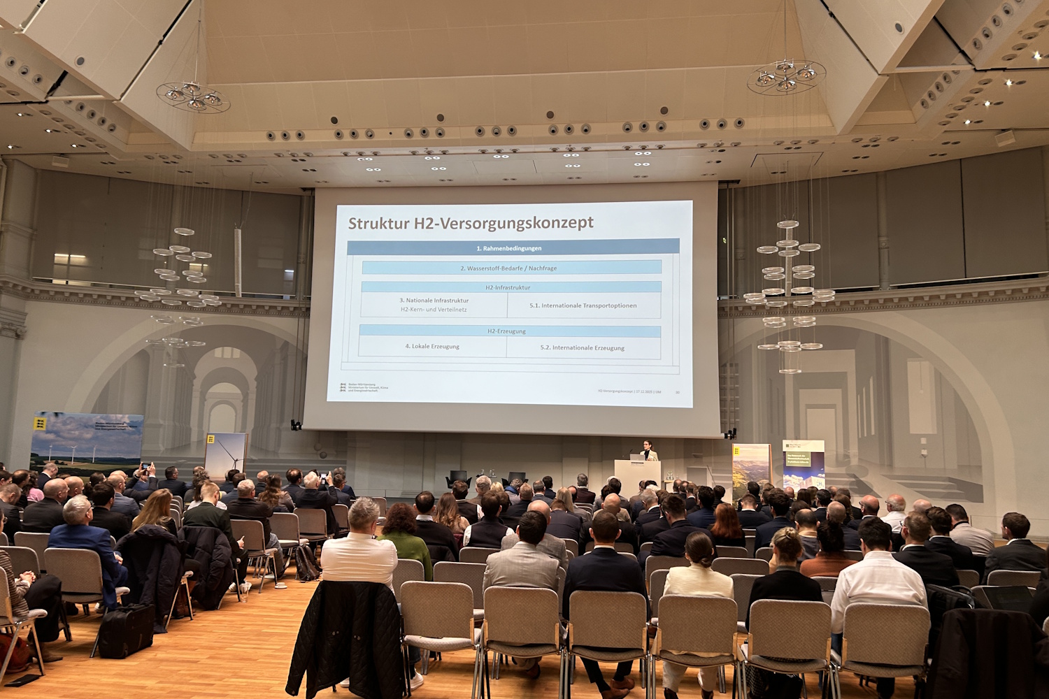 Rund 200 Personen sitzen in Stuhlreihen in einem großen Saal und blicken nach Vorne, wo großflächig eine Präsentation gezeigt wird.