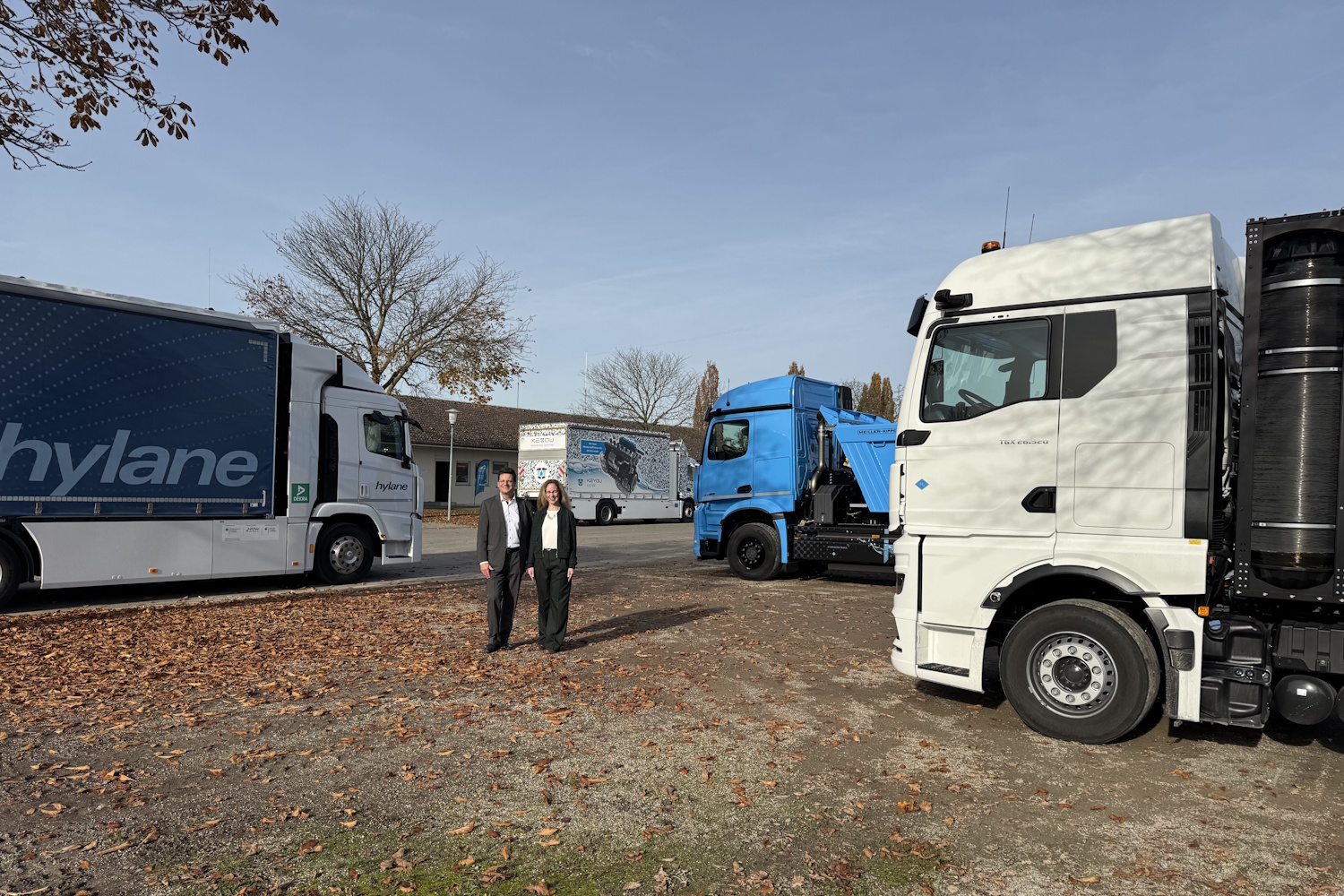 Zwischen drei großen Lkw stehen zwei Personen auf freier Fläche.