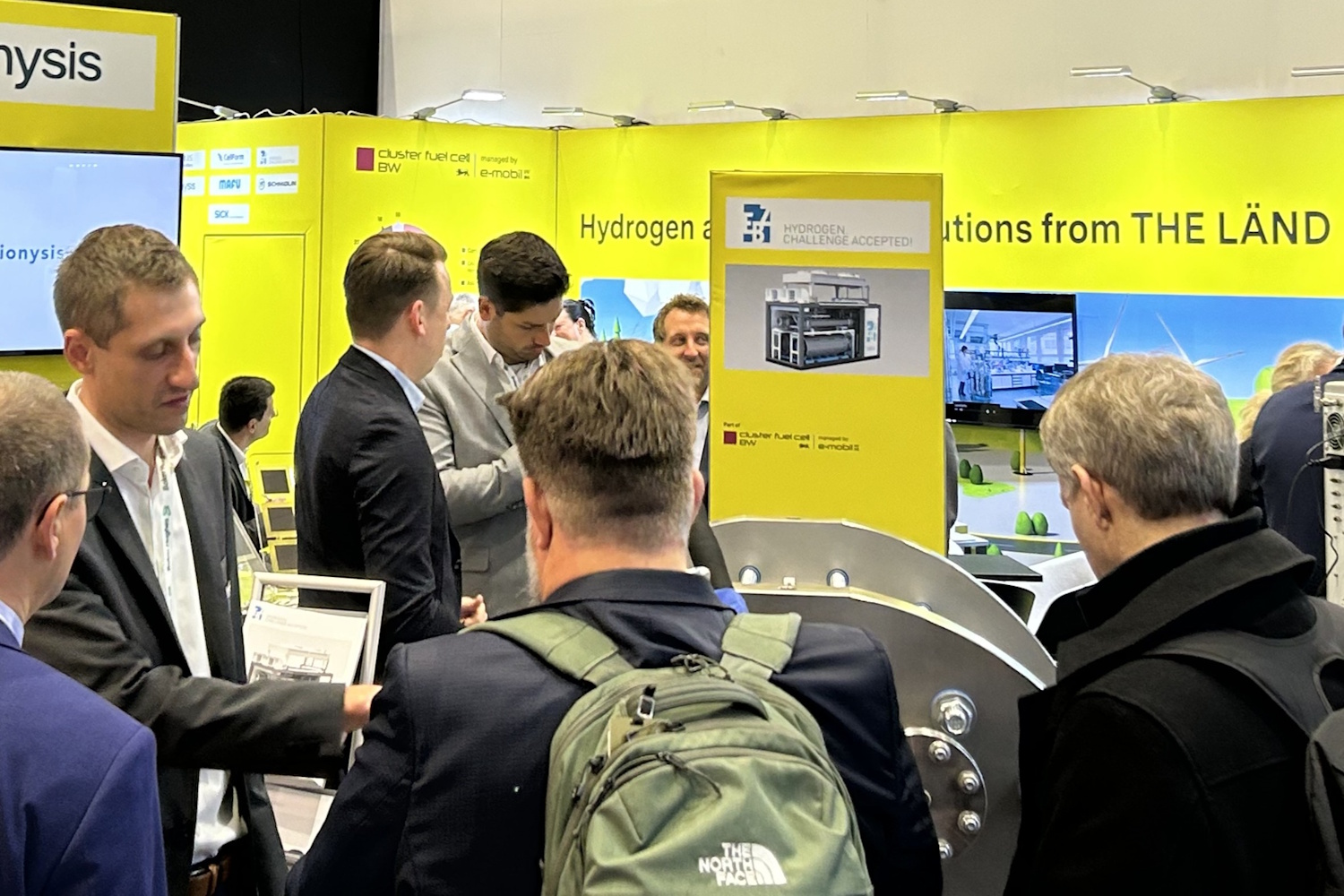 Eine Gruppe Menschen steht auf einem gelben Messestand und lässt sich etwas erklären.