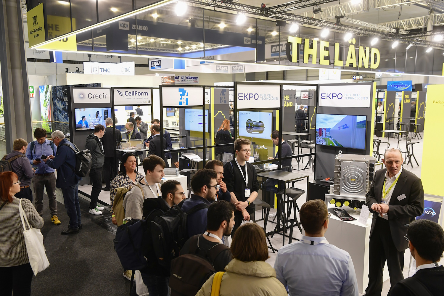 Eine Gruppe Menschen steht vor dem Messestand von THE LÄND. Man sieht viele Logos von ausstellenden Unternehmen an ihren kleinen Messeständen.